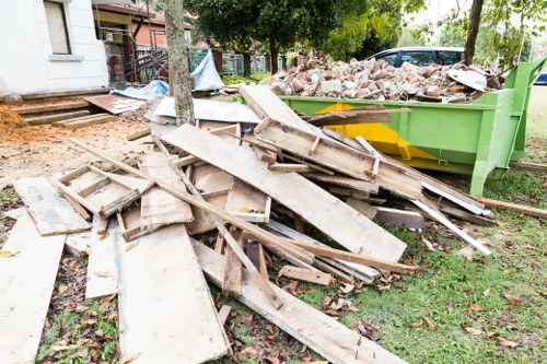 A pile of discarded wooden decking boards and planks, some weathered and uneven, rests on a grassy area near a large green skip filled with mixed construction debris, including bricks and broken materials, in an outdoor residential setting. The background features trees, a pathway, and parts of a building, suggesting an ongoing rubbish removal or garden clearance service provided by Office Clearance Kingston in Kingston upon Thames. The scene is illuminated by natural daylight, highlighting the textures of the wood, brick rubble, and the surrounding environment, with a focus on waste removal for property clearance in the local area.