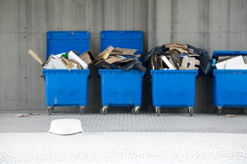 A row of five blue wheelie bins positioned on a paved surface against a grey concrete wall. The first two bins from the left are filled with mixed waste, including cardboard, paper, and plastic debris, with some of the items partially protruding over the rim. The third bin appears to be overflowing with waste, with a black plastic rubbish bag on top spilling out items such as cardboard boxes and paper. The fourth bin contains similar mixed rubbish, slightly less overflowing, and the fifth bin is partially obscured with additional waste visible. In the foreground, a white plastic container, possibly a discarded lid or small bin, rests on the ground. The scene is outdoors, likely in an urban or commercial area, with the company's rubbish removal service in Kingston evident in the context of the image.