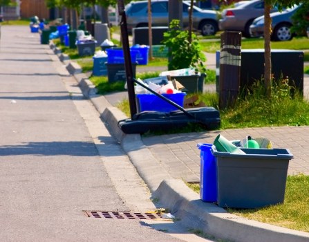 A clear urban sidewalk in a residential area with various bins and containers placed along the curbside for rubbish collection. In the foreground, a large gray plastic storage bin contains green and white plastic bottles, positioned on a grassy verge beside the pavement. To its right, a smaller blue bin is visible. Further down the sidewalk, several blue and black skip bins and boxes are arranged along the edge of the pavement, some partially filled with mixed waste. A yellow-handled broom is leaning against one of the bins, indicating ongoing or recent cleaning activity. Behind these containers, a row of young trees with green foliage lines the street, and parked cars can be seen in the background, along with a curb and a drainage grate inset into the pavement. The overall scene depicts a typical rubbish collection or clearance setup near a residential property, consistent with services offered by Office Clearance Kingston, serving Kingston and nearby areas.
