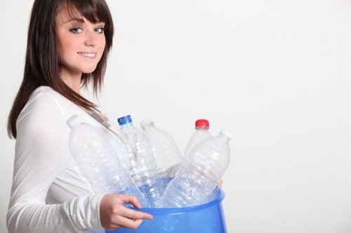 A young woman with shoulder-length brown hair and fair skin, wearing a white long-sleeved top, is holding a blue plastic bucket filled with empty clear plastic bottles that have varying cap colors, including red, blue, and white. She stands against a plain white background, smiling gently while looking slightly to her right. The bottles have textured surfaces and are of different shapes and sizes, with some featuring ridges and others smooth. The scene appears well-lit, emphasizing the transparency and glossy finish of the plastic bottles. The image suggests waste collection or rubbish disposal, relevant to a rubbish removal service in Kingston, with the woman presenting an organized collection of recyclable plastic packaging, aligned with the services offered by Office Clearance Kingston in the local area.