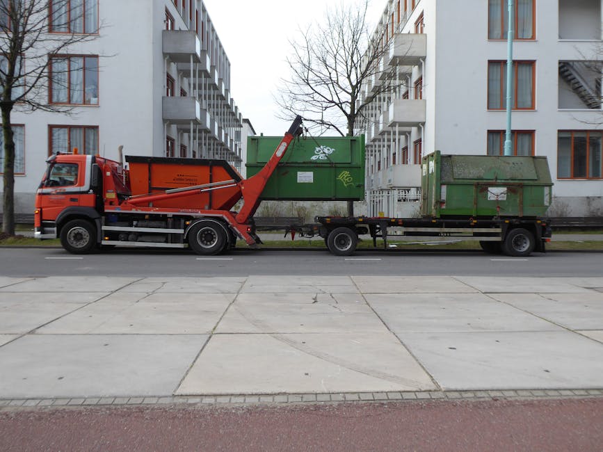 A male waste collection worker wearing an orange and dark grey high-visibility uniform is standing beside a large, white and yellow rubbish collection vehicle at dusk or night, with orange and red sun